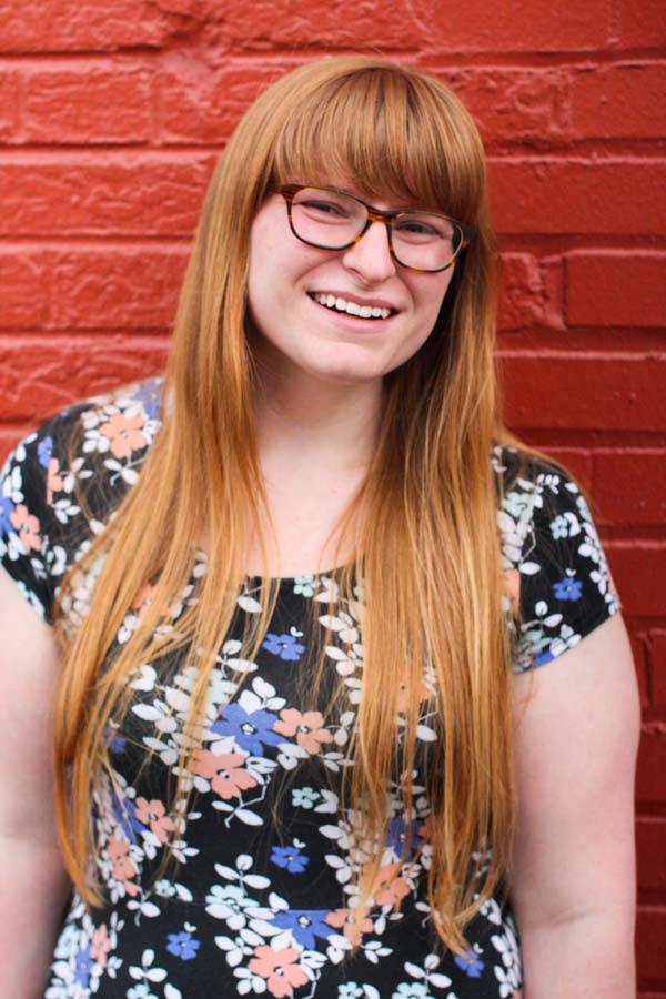 Portrait of Katie standing in front of a red brick wall.